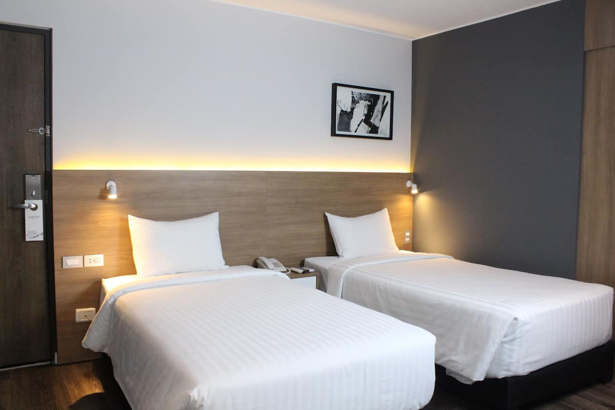 Een hotelkamer met twee bedden, een houten hoofdbord, een nachtkastje met telefoon en een ingelijste foto aan de muur.