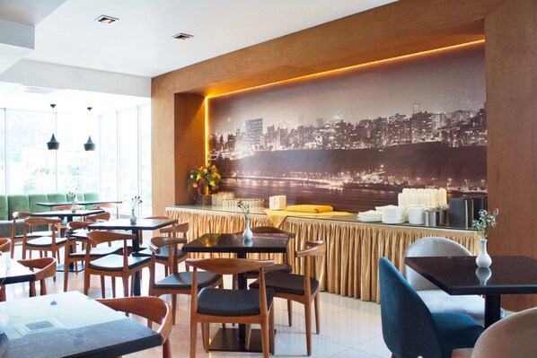 Un restaurante con varias mesas y sillas, un mural de la ciudad en la pared y una zona de bar con mostrador amarillo.