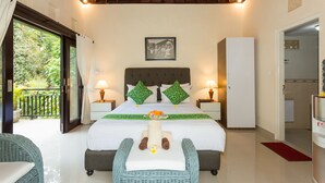 A hotel room with a bed, two nightstands, a wicker chair, a red rug, and a balcony with a view of greenery. - Villa Bakti Ubud (Ubud)