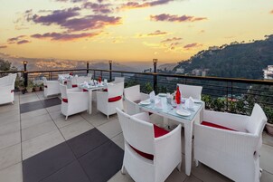 Una terraza con muebles de mimbre blancos y mesas de vidrio, con vista a un paisaje montañoso al atardecer.