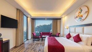 A hotel room with a large bed, a TV, a desk, and a balcony with a view of a hill. - Snow Valley Resorts (Shimla)