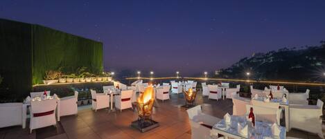 Un área de comedor al aire libre con mesas y sillas, una chimenea y vistas al cielo nocturno.