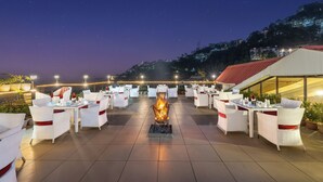 A rooftop terrace with tables and chairs arranged for dining, a central fire feature, and a view of a hillside with buildings. - Snow Valley Resorts (Shimla)