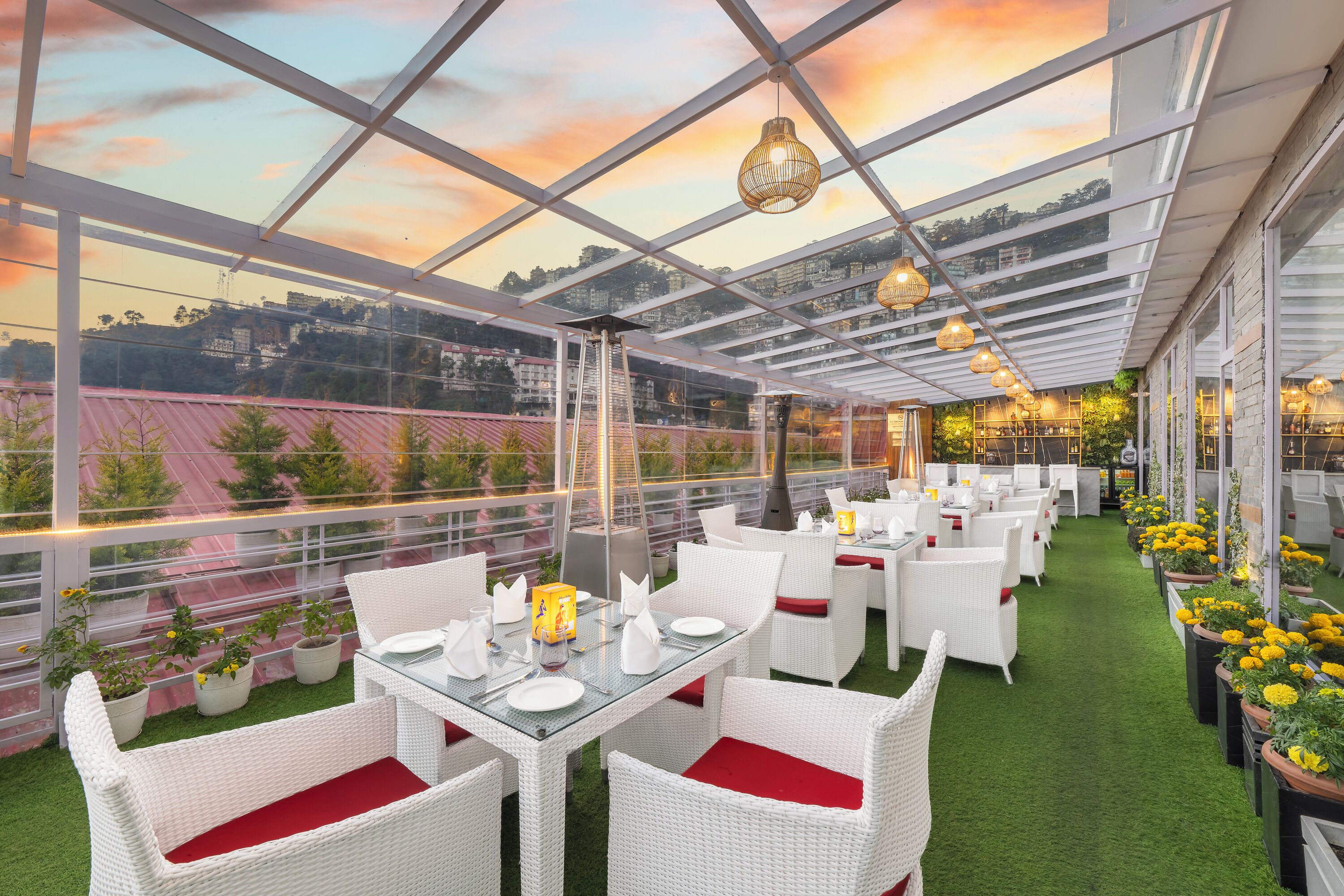 An outdoor restaurant with white wicker furniture and red cushions, set against a backdrop of a sunset sky and cityscape.