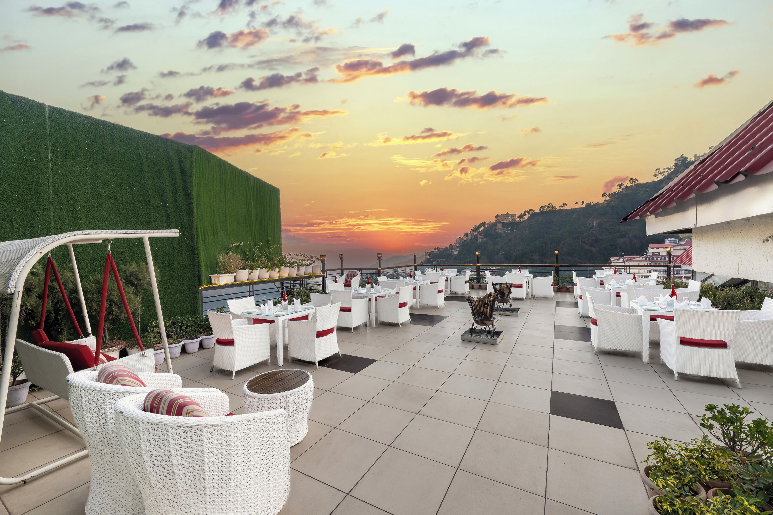 Terraço no telhado com móveis de vime branco, almofadas vermelhas e vista para um pôr do sol sobre uma paisagem montanhosa.