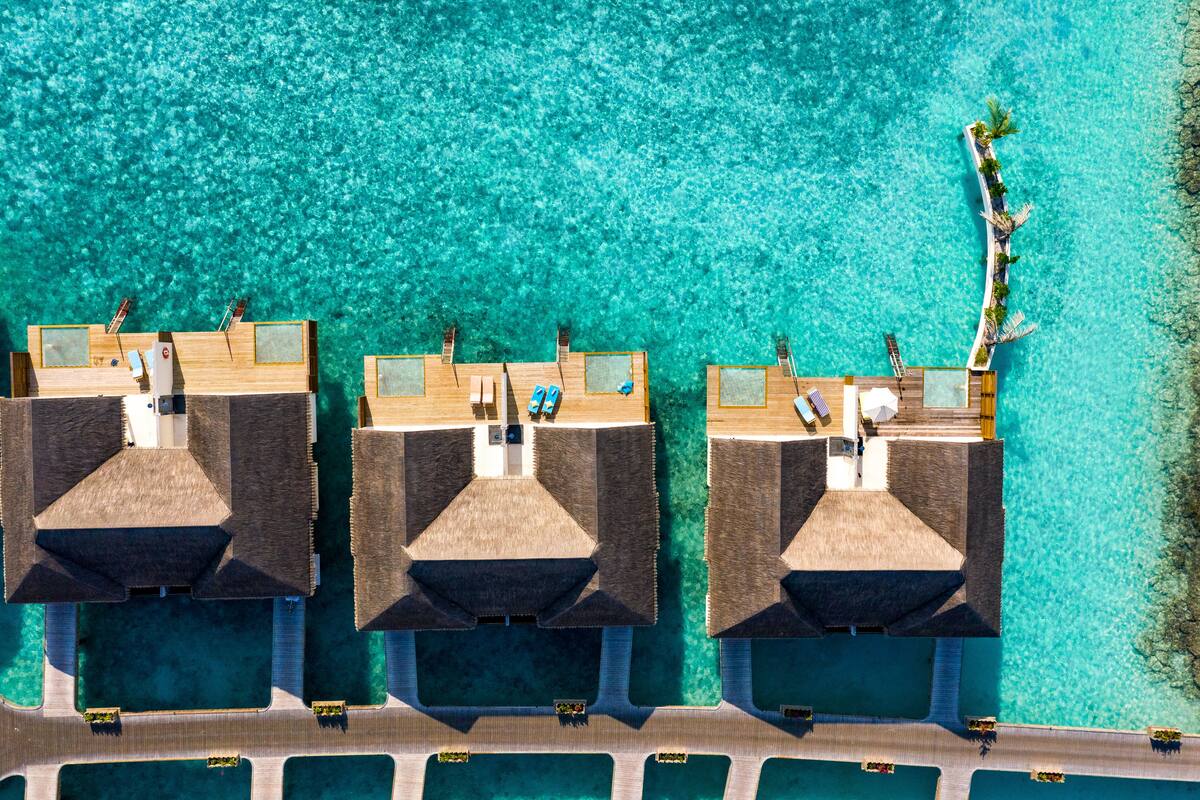 An aerial view of a resort with three overwater bungalows connected by a wooden walkway, surrounded by clear turquoise water.