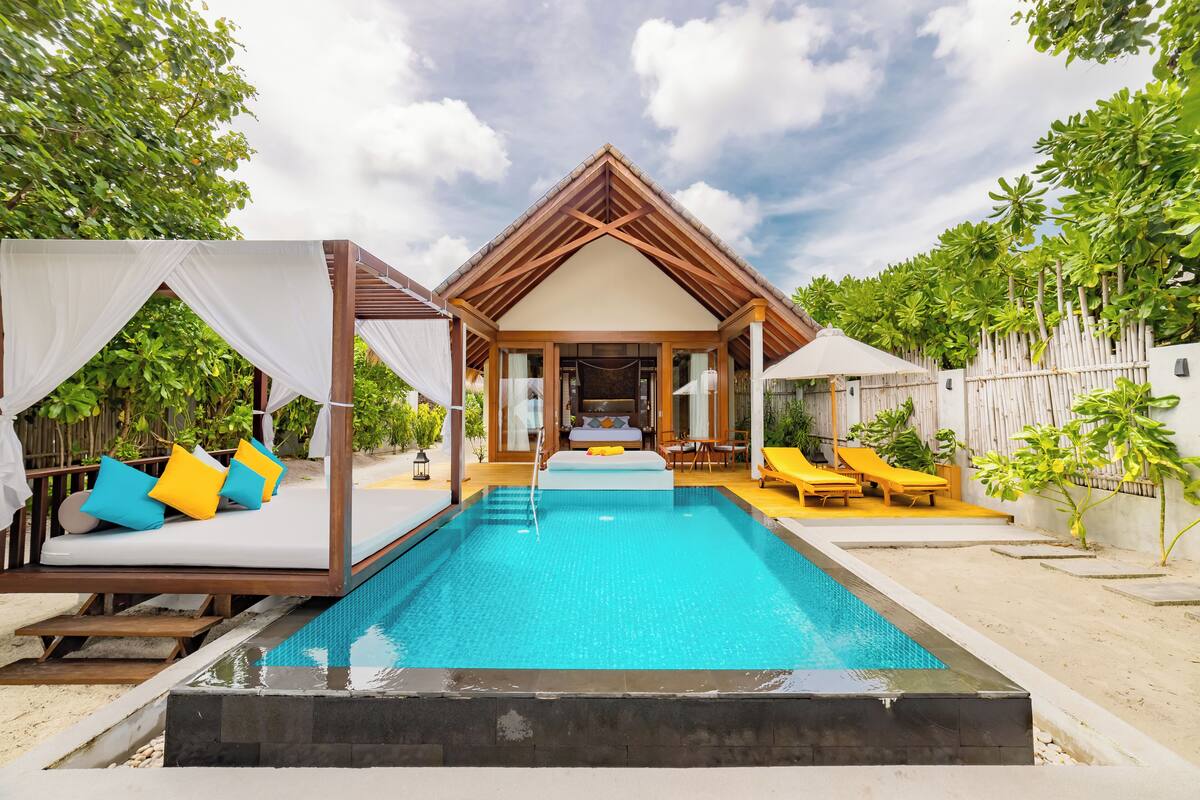 A poolside area with a wooden deck, a covered seating area with cushions, and a view of a building with a triangular roof.