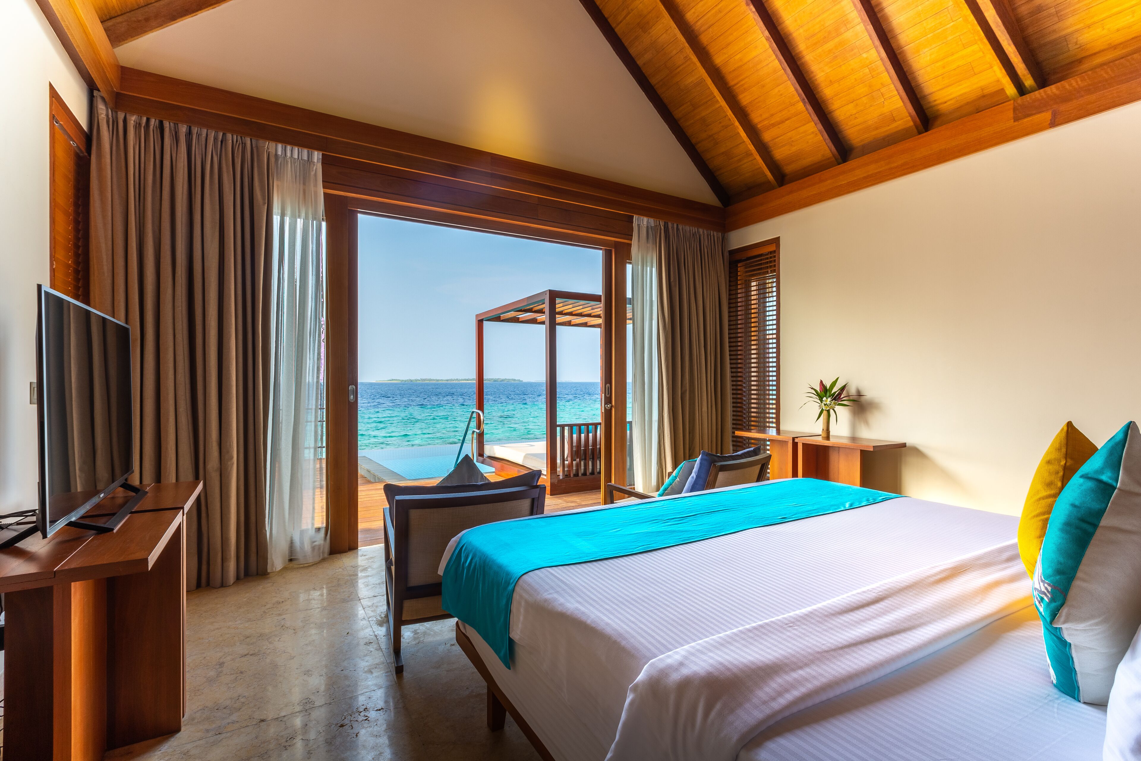 A hotel room with a large bed, a desk with a TV, a chair, and a view of the sea through large windows.
