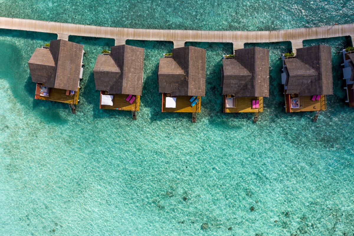 An aerial view of overwater bungalows with thatched roofs, connected by a wooden walkway.