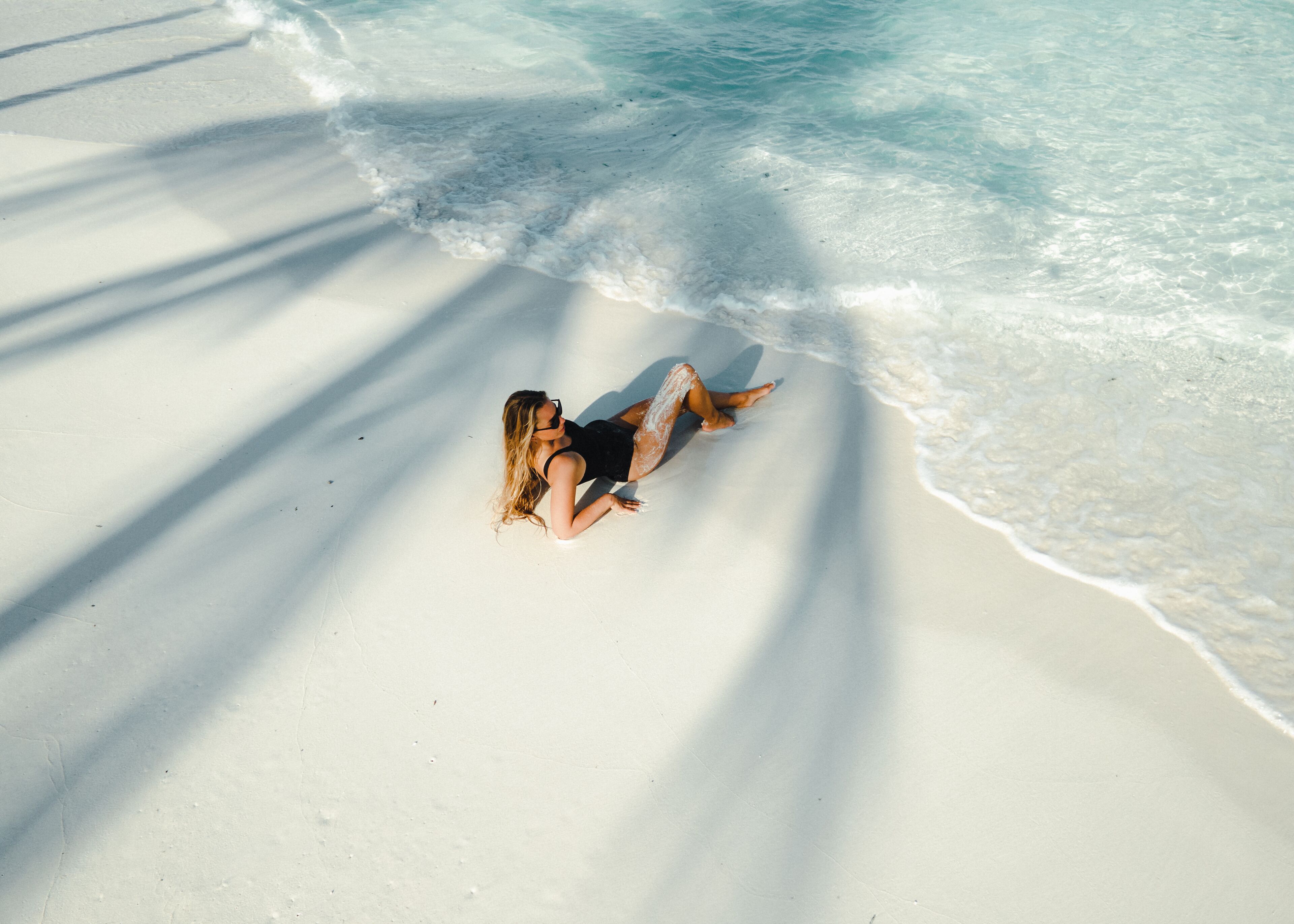 A person lying on a sandy beach with clear water and shadows cast by palm trees.