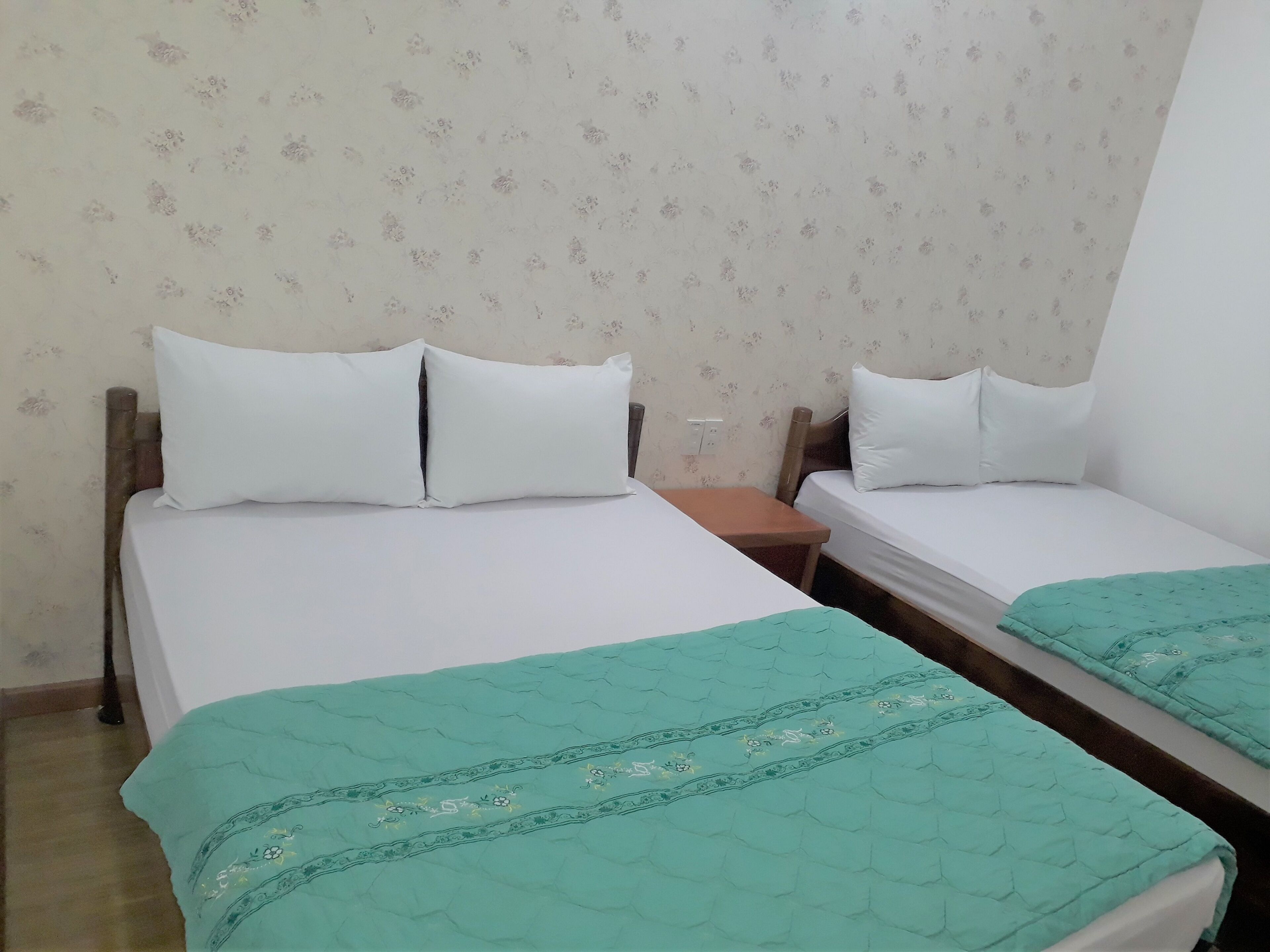 Une chambre avec deux lits simples, chacun doté de draps blancs et d’une couette verte. La chambre présente une disposition simple, avec une tête de lit en bois et un mur sobre orné d’un motif floral.