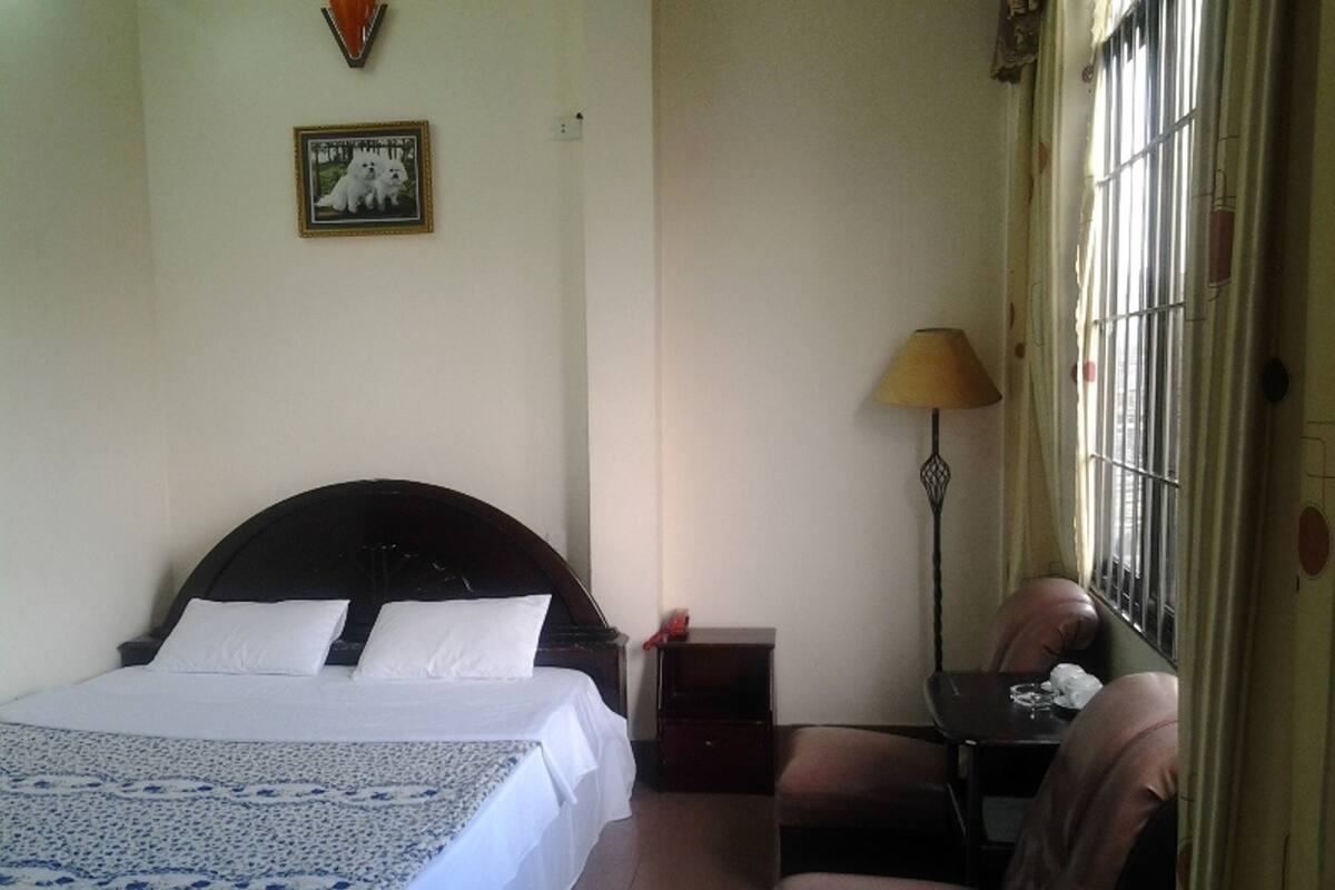 A bedroom with a bed, a chair, a nightstand, a lamp, and a window with bars.