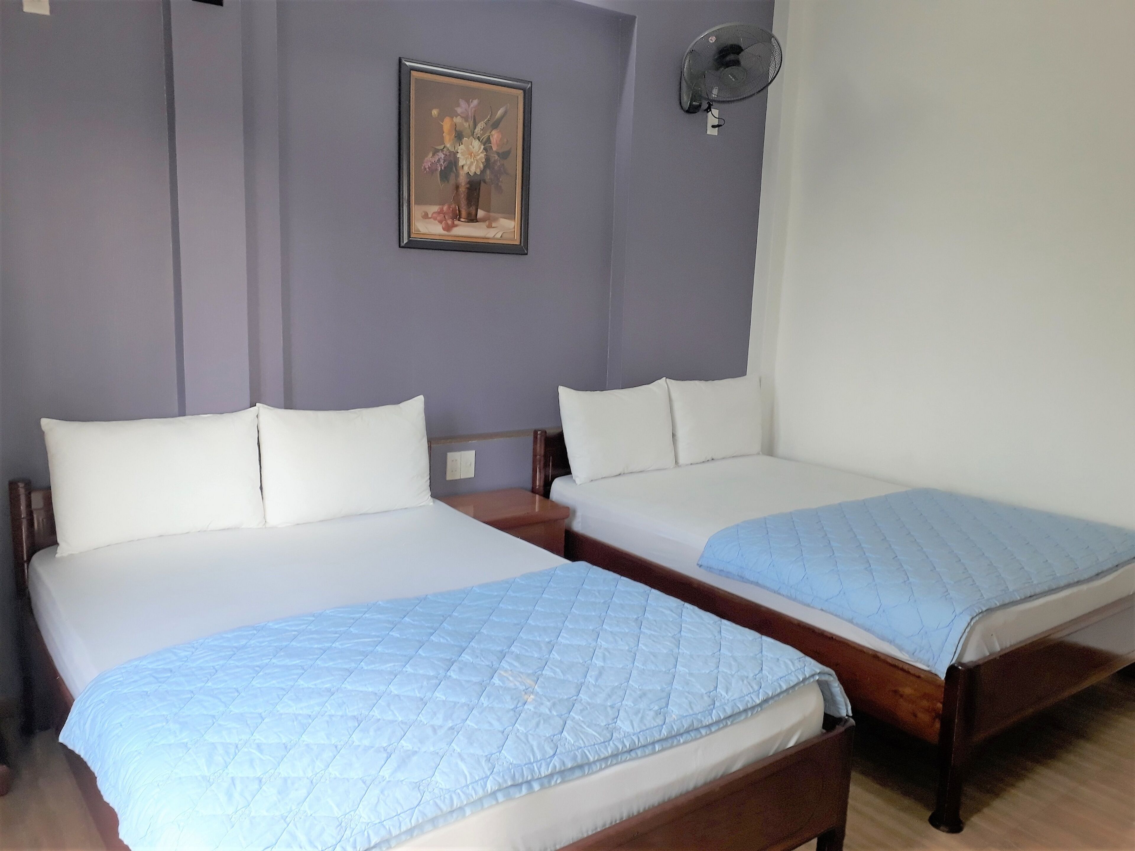 A hotel room with two beds, a nightstand, a wall-mounted fan, and a framed floral painting.