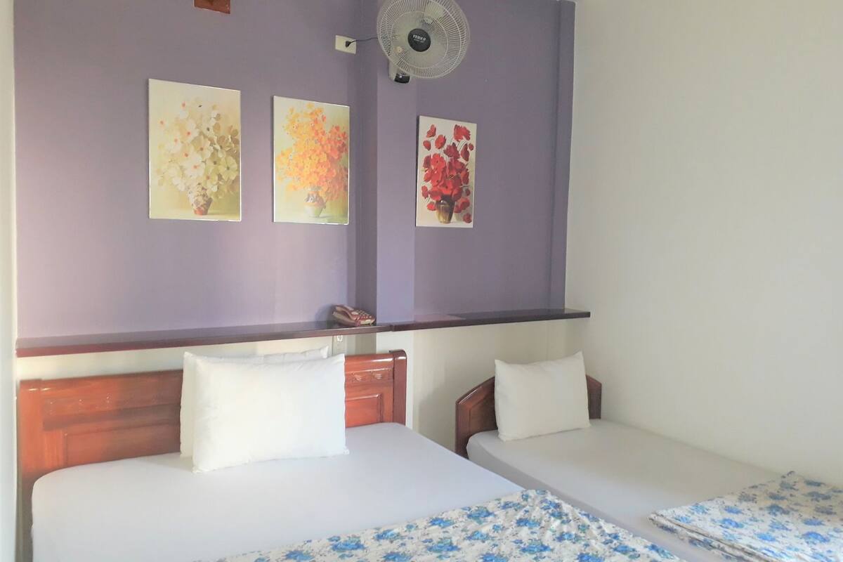 A bedroom with two beds, a wall-mounted fan, and three framed floral paintings.