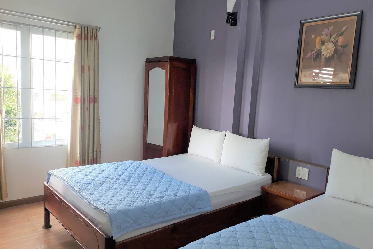 A hotel room with two beds, a large window with curtains, a mirror, and a framed floral painting on the wall.