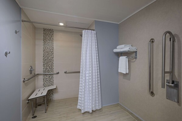 Ducha accesible en silla de ruedas con cortina blanca, barra de apoyo y repisa con toallas.