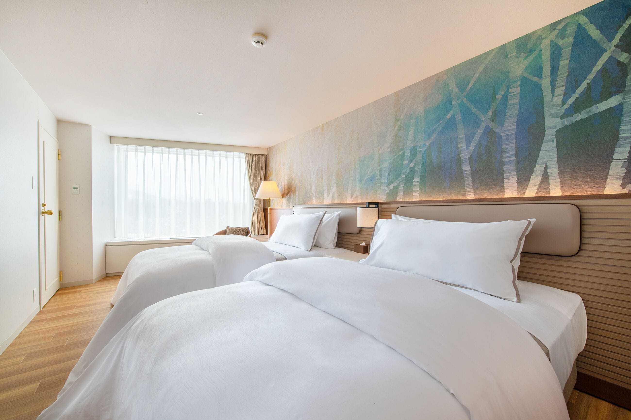 A hotel room with two beds, a large window with curtains, a painting on the wall, and a lamp on the nightstand.
