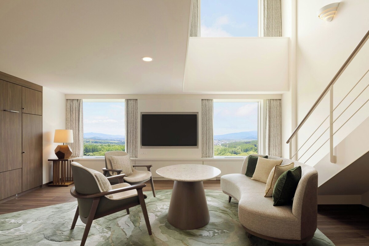 A spacious living room with a large flat-screen TV, comfortable seating, a staircase, and a view of the outdoors.