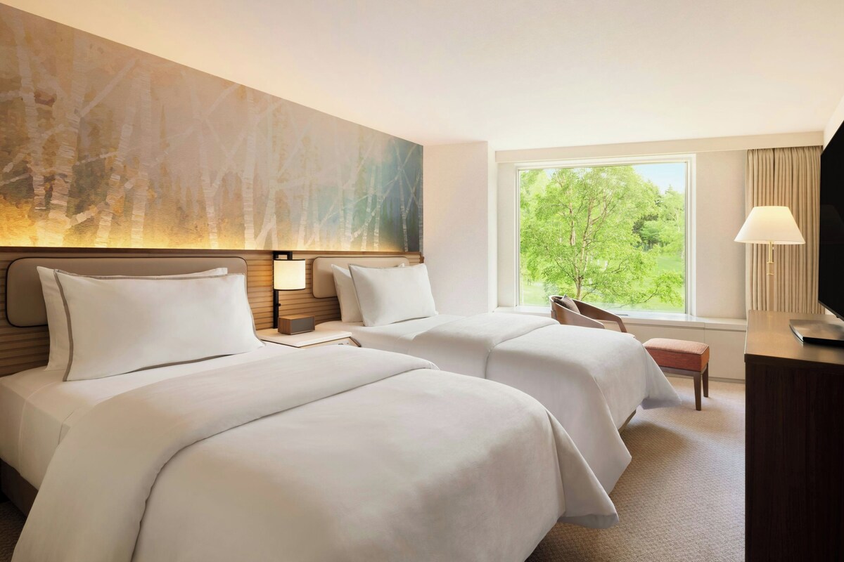 A hotel room with two beds, a large window with a view of greenery, a desk with a lamp, and a painting on the wall.