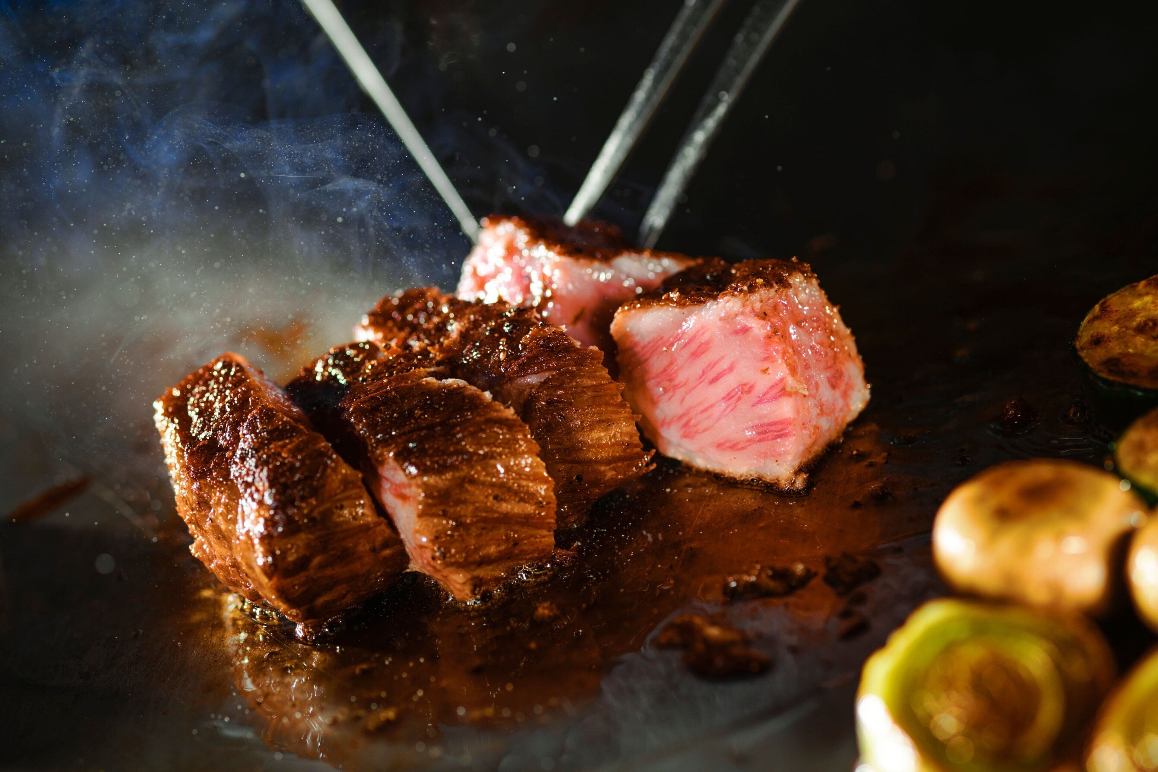 A close-up of grilled meat with chopsticks, steam, and roasted potatoes.