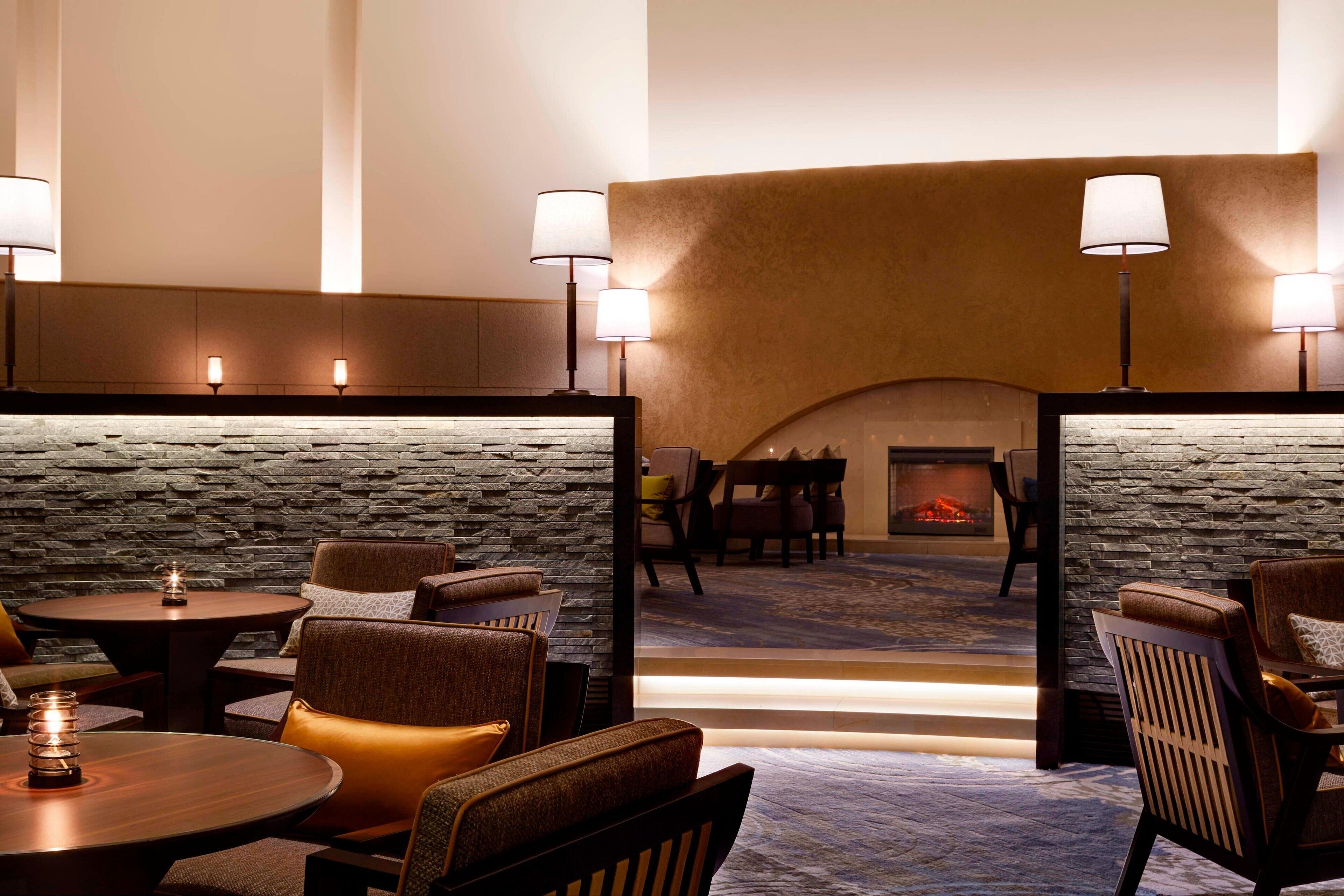 A cozy lounge area with round tables and cushioned chairs, a stone wall, and a fireplace in the background.