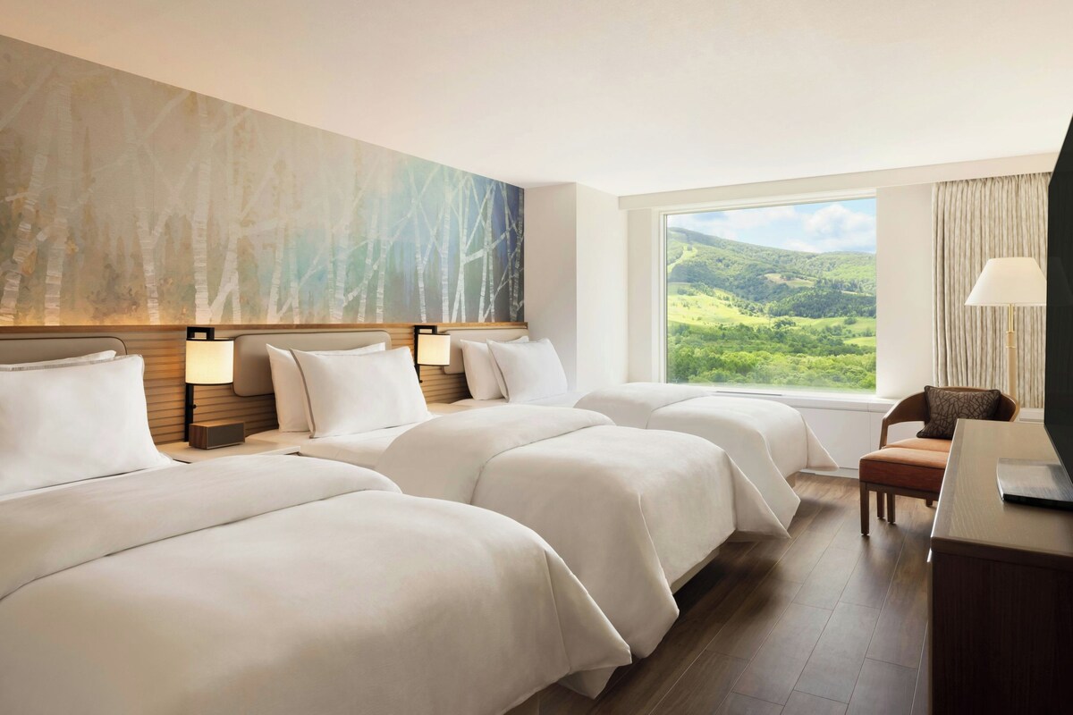 A hotel room with three beds, a large window with a view of a green landscape, and a desk with a lamp.