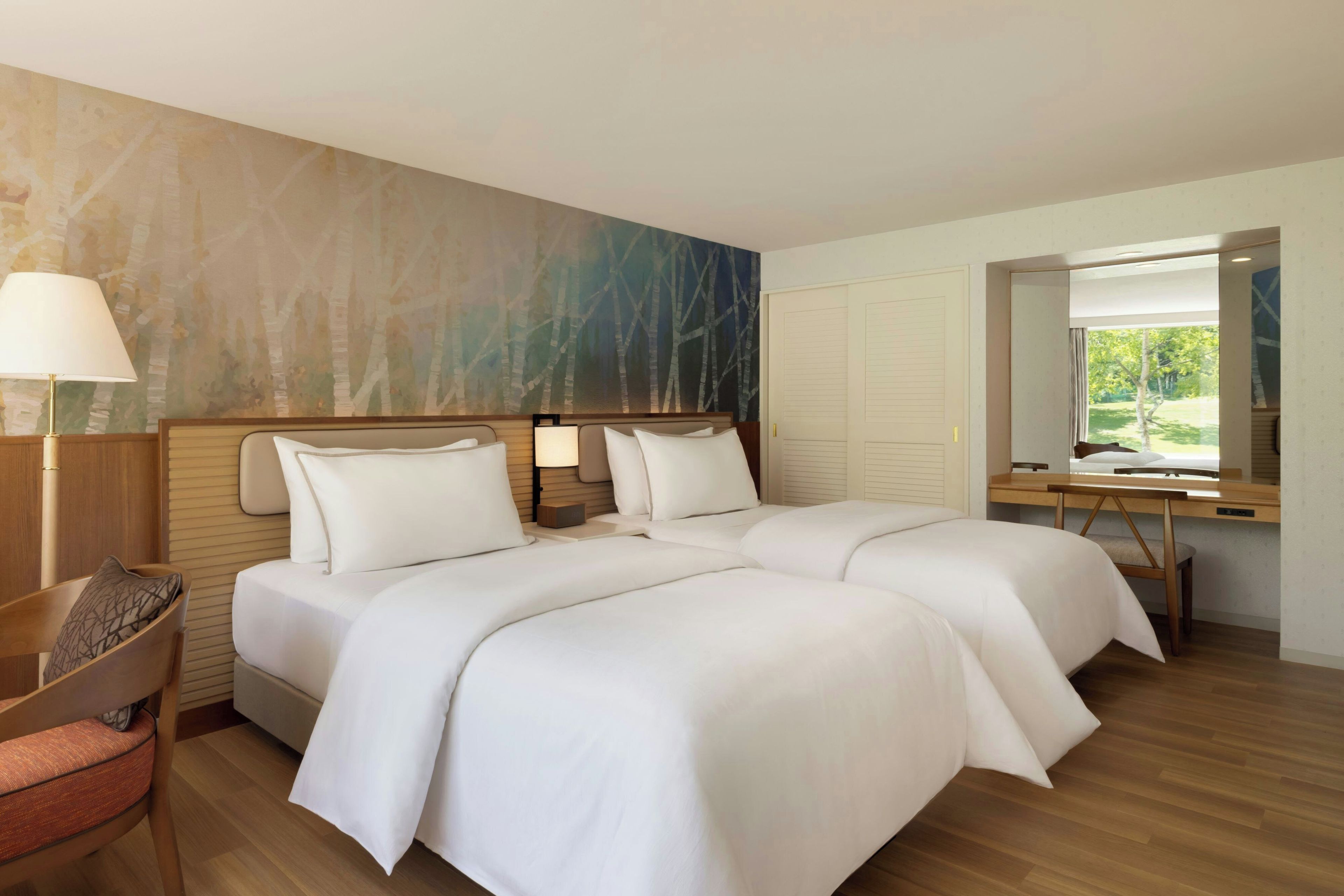 A hotel room with two beds, a desk, a chair, a lamp, and a large window with a view of greenery.