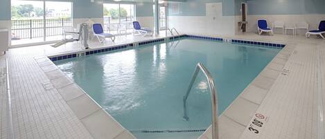 Une piscine à l’eau d’un bleu cristallin, entourée de carrelages blancs au sol et sur les murs, avec quelques chaises longues et une fenêtre donnant sur l’extérieur.