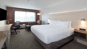 Ein Hotelzimmer mit einem großen Bett, einem Schreibtisch mit Stuhl, einem Fernseher und einem Fenster mit Blick auf die Stadt.