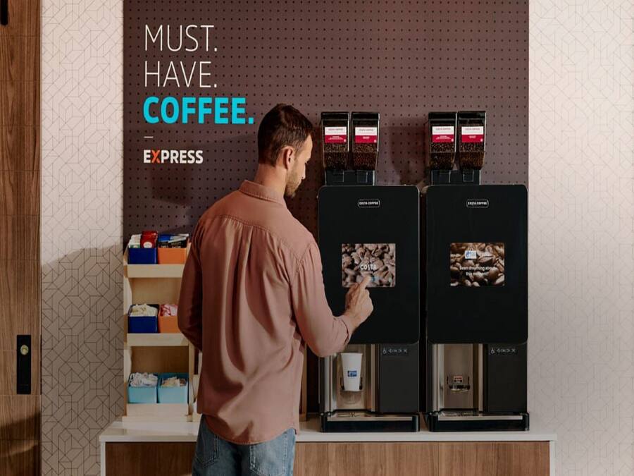 A man in a brown shirt interacting with a coffee machine, with a sign above stating "MUST. HAVE. COFFEE."