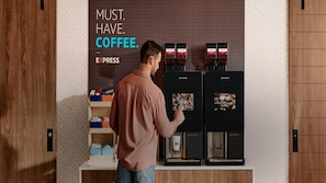 A man in a brown shirt interacting with a coffee machine, with a sign above stating "MUST. HAVE. COFFEE."