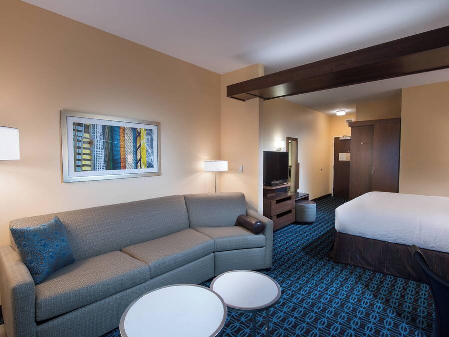 Habitación de hotel con sofá, dos mesitas redondas, cama, televisión e imagen en el marco con vista panorámica de la ciudad.