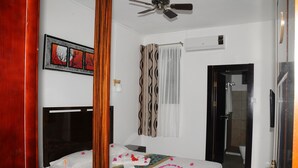 A hotel room with a bed, a bedside table, a nightstand, a ceiling fan, an air conditioning unit, a shower area, and a door leading to another room. - Le Flamboyant Hotel (Rodrigues Island)