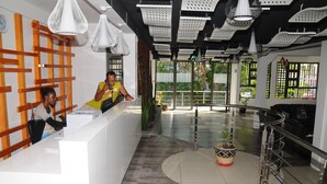 A reception area with a white counter, modern pendant lighting, and a view of the outdoors through large windows. - Le Flamboyant Hotel (Rodrigues Island)