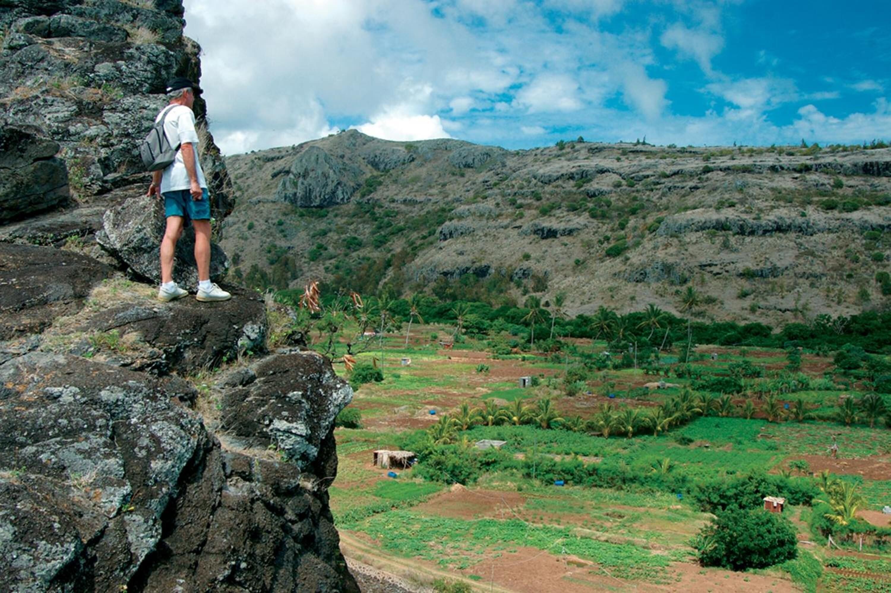A person standing on a rocky outcrop overlooking a valley with scattered buildings and greenery.
