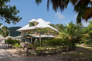 Une maison à deux étages, dotée d’un toit en pente, entourée d’une végétation tropicale et baignée sous un ciel dégagé.