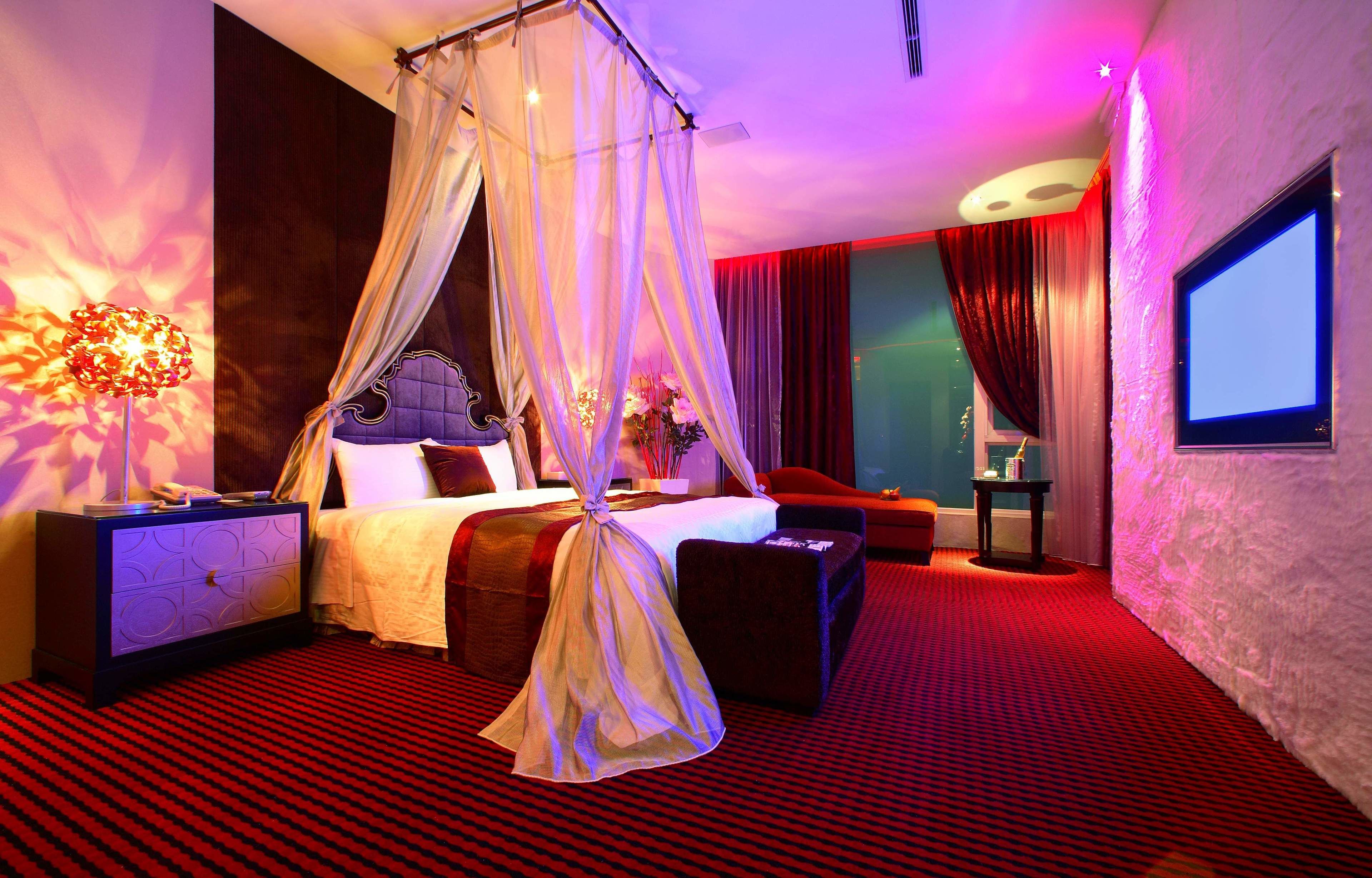 A hotel room with a four-poster bed, a flat-screen TV, a nightstand, a bedside table, and a red carpeted floor.