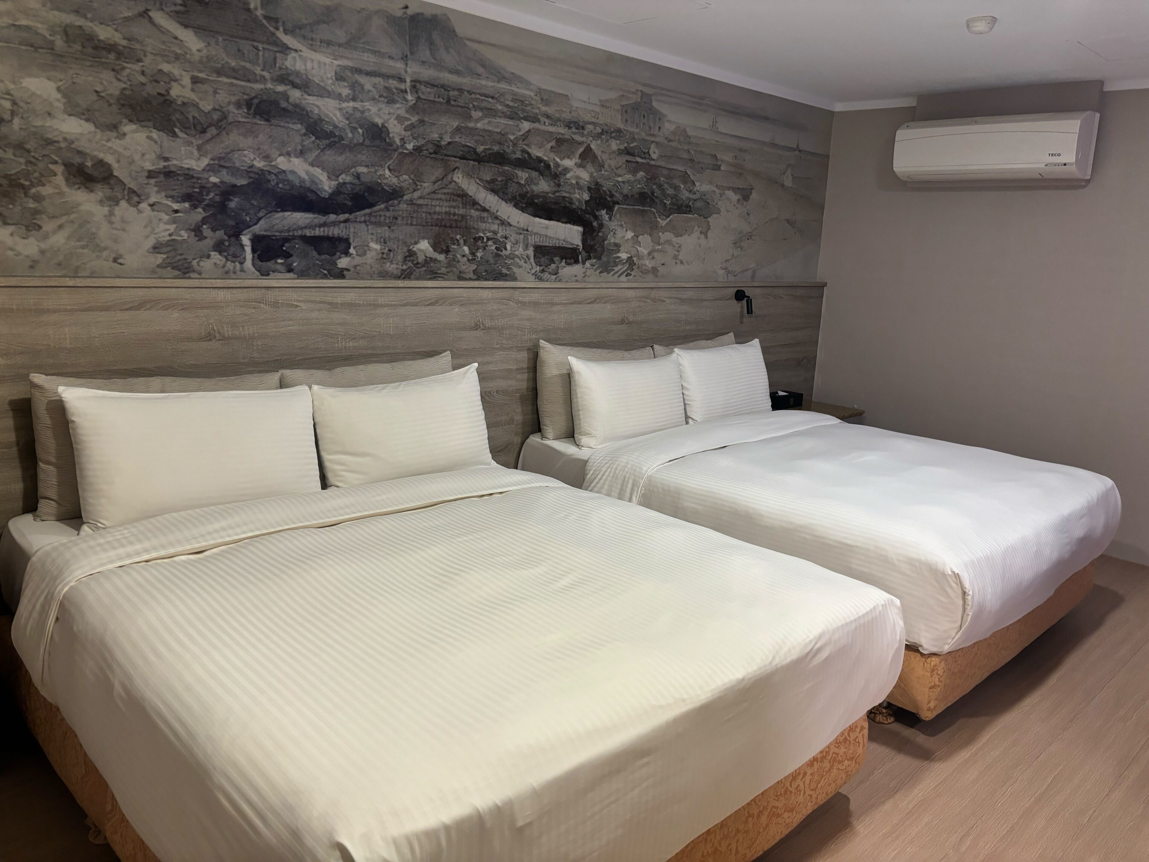 A hotel room with two beds, a large wall mural, and a ceiling-mounted air conditioner.