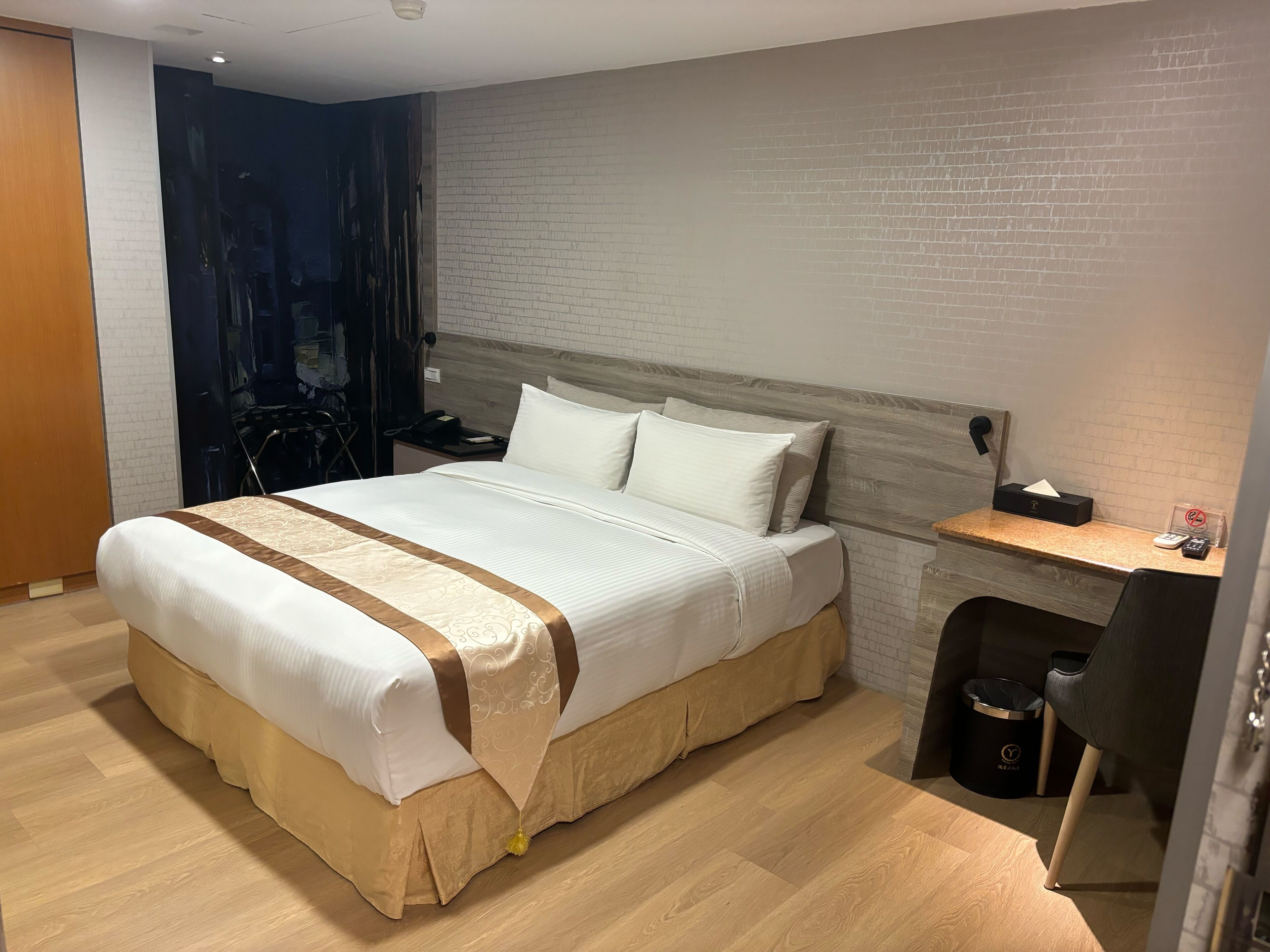 A hotel room with a large bed, a wooden headboard, a nightstand with a phone and a tissue box, a desk with a chair, and a trash bin.