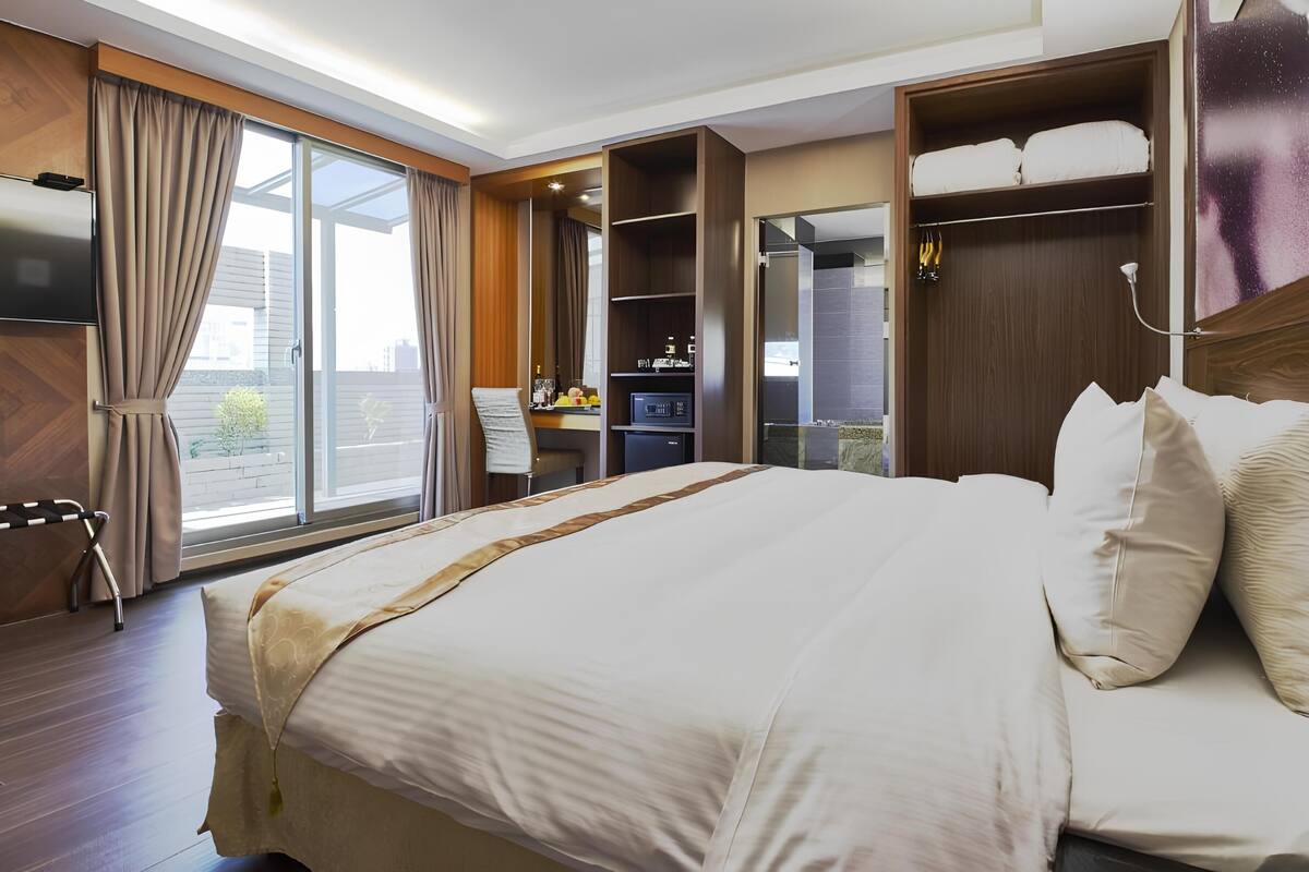 A modern hotel room with a large bed, a desk with a chair, a wardrobe, and a TV.