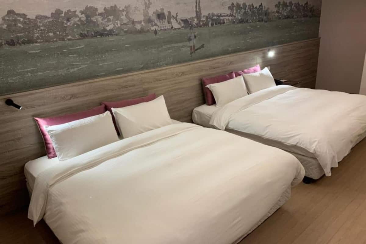 A hotel room with two beds, each with white bedding and pink pillows, a large painting on the wall depicting a landscape, and wooden flooring.