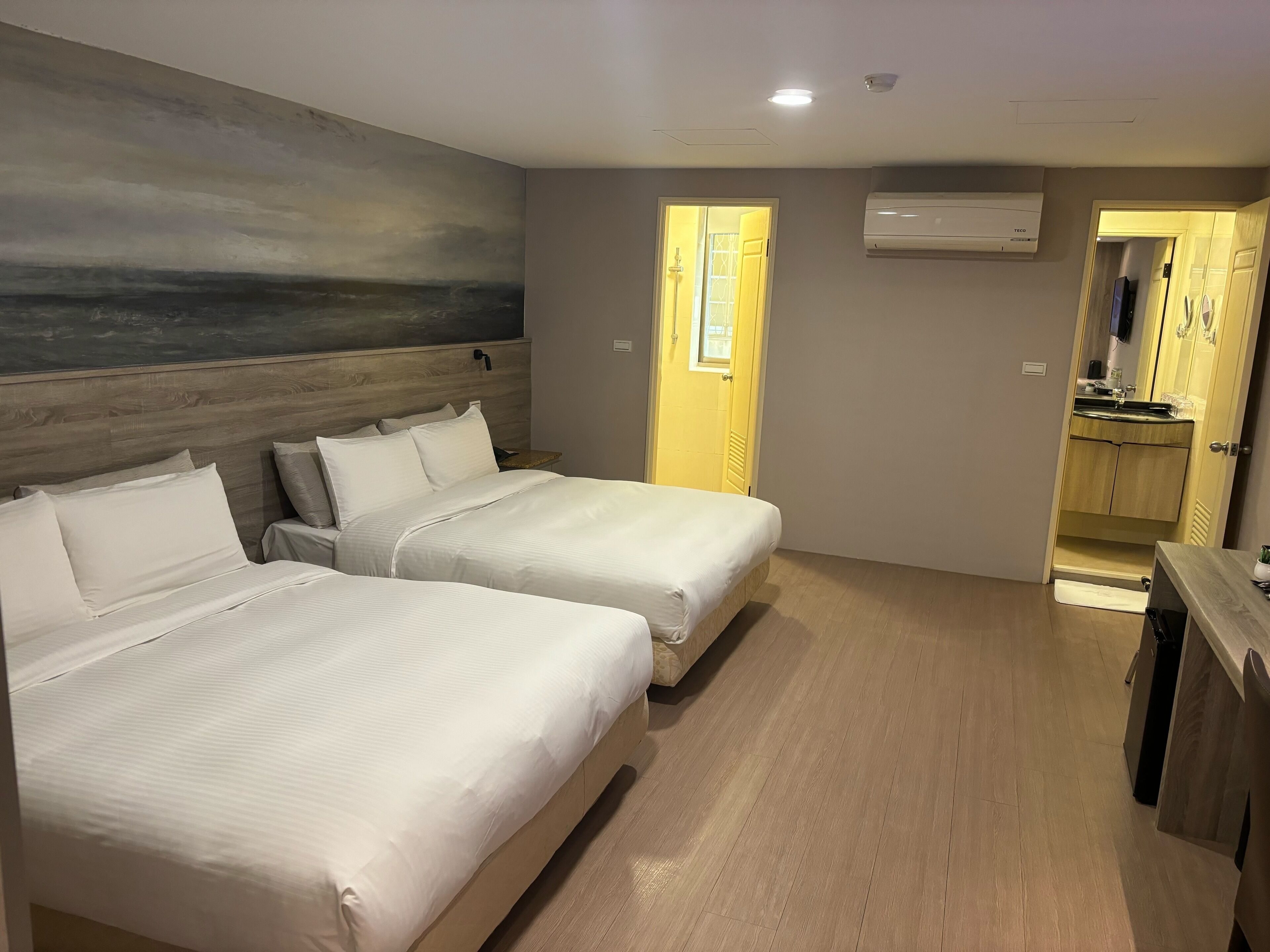 Habitación de hotel con dos camas, cabecero de madera, un cuadro en la pared, puerta de baño y una zona de kitchenette.