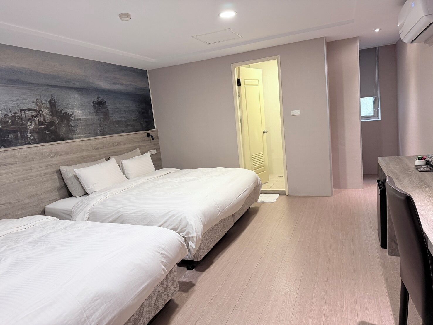 A hotel room with two beds, a wooden headboard, a painting on the wall, a desk with a chair, and a door leading to another room.