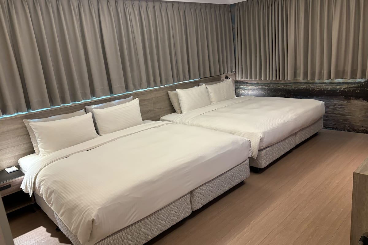A modern hotel room with two beds, a wooden headboard, and a view of a stone wall.