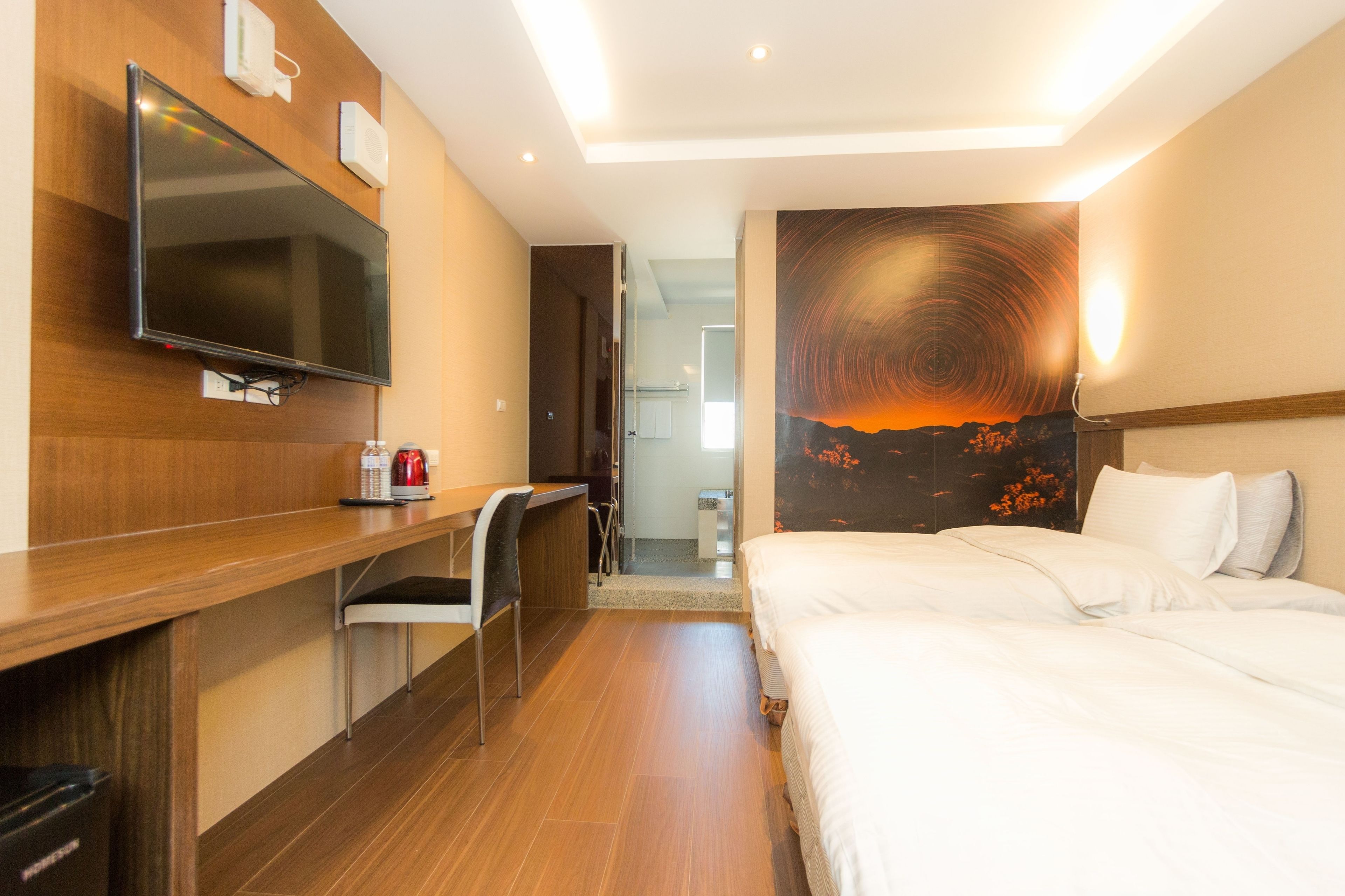 A hotel room with a bed, a desk with a chair, a television, and a painting of a sunset.