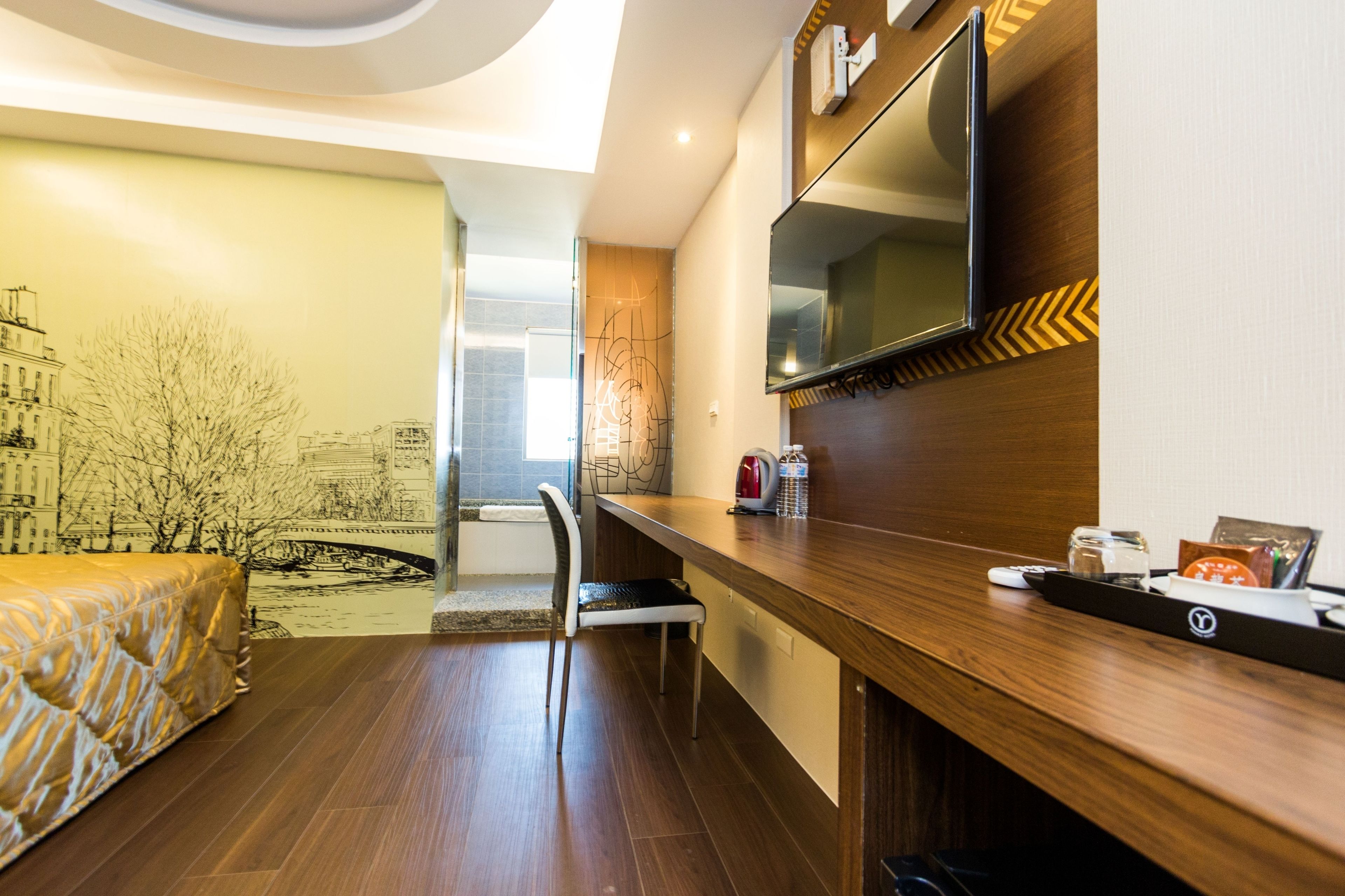 Una habitación de hotel moderna con un escritorio de madera, una silla, una cama, un televisor y un mural en la pared con vista a un paisaje urbano.