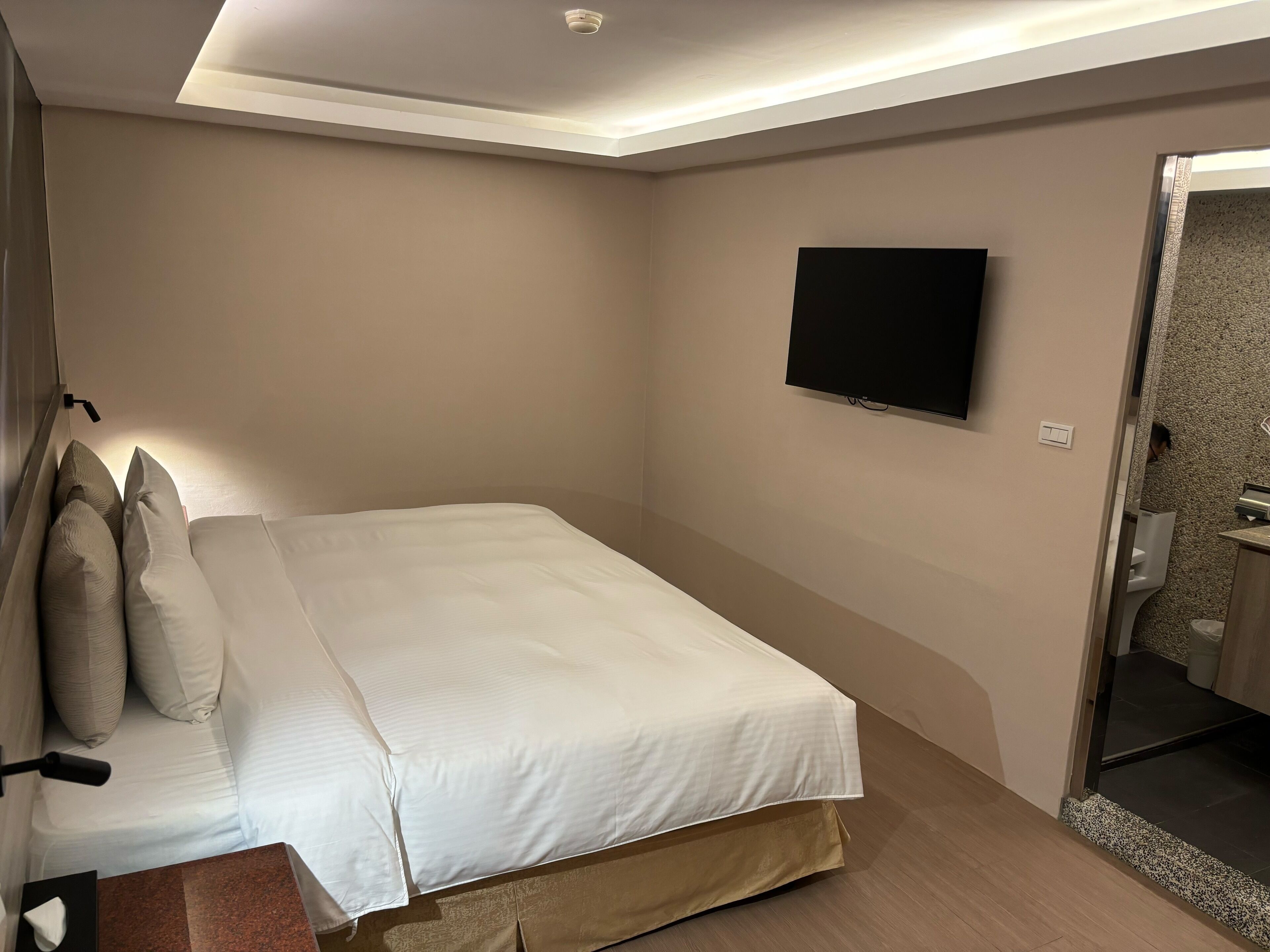 A hotel room with a bed, a TV mounted on the wall, and a bathroom visible through an open door.