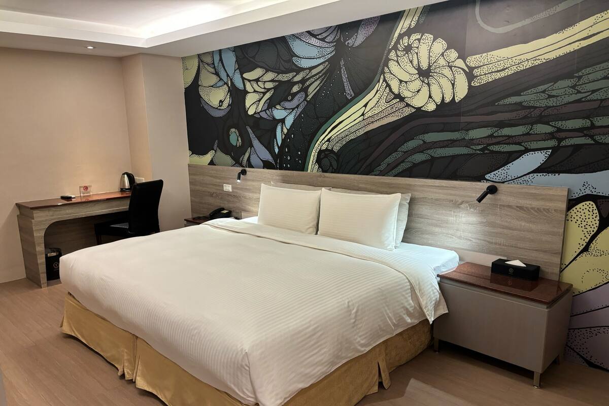 A hotel room with a large bed, a desk, a chair, and a nightstand.