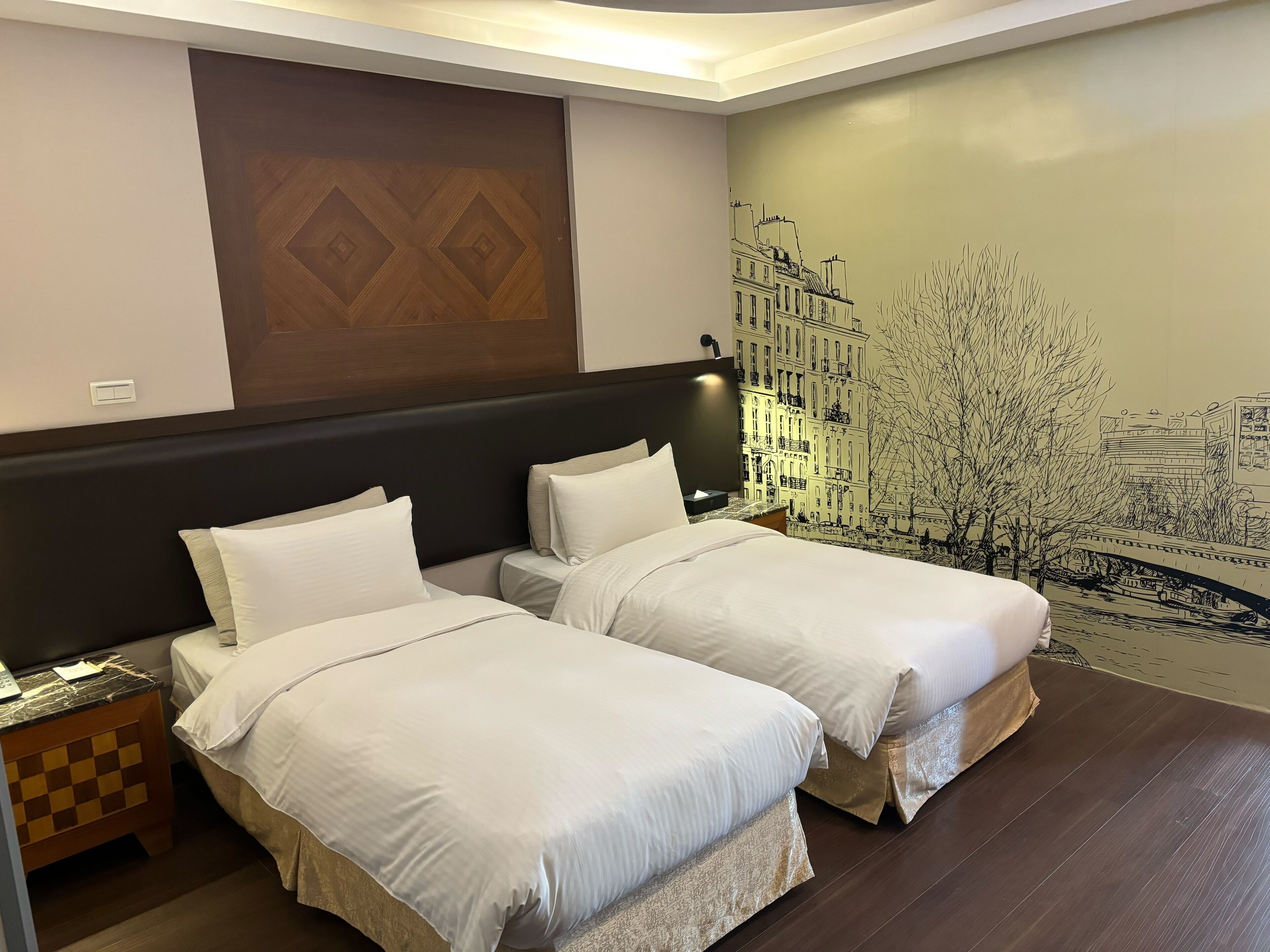Une chambre d’hôtel avec deux lits simples, une tête de lit en bois, une fresque murale, une table de chevet et une lampe.