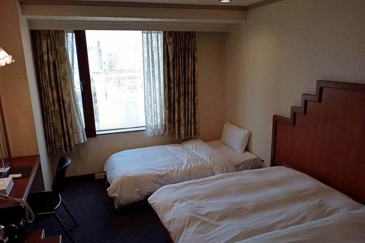 Une chambre d’hôtel avec deux lits, un bureau et une chaise. Il y a une fenêtre avec des rideaux, et on aperçoit un escalier au fond.